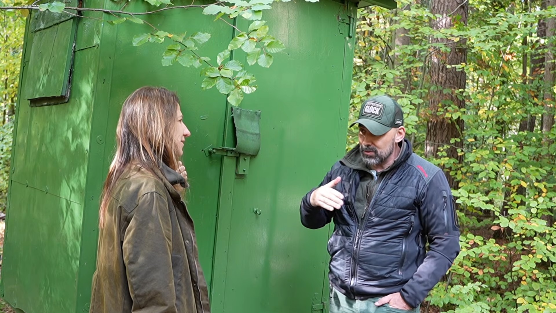 Vedrana Miholić and Martin Pluschke talking beside a green hunting shelter in a forest near Nuremberg.